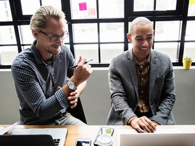 Two men sitting at a table looking at a computer and talking